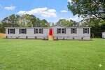 A single-story gray manufactured home with a bright red front door and small porch steps sits on a large, well-maintained green lawn with trees and a clear sky in the background.