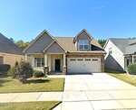 A two-story suburban house with beige siding, stone accents, a covered front porch, and a double garage. The house has a well-maintained front lawn and sits between two similar homes under a clear blue sky.