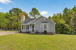 A single-story gray house with white trim sits on a large, grassy lawn. There are a few shrubs by the covered front porch, and the house is surrounded by trees under a clear blue sky.