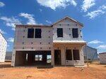 A two-story house under construction with exterior sheathing and window openings but no doors or windows installed; the surrounding ground is bare dirt under a partly cloudy sky.