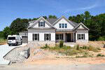 A newly constructed two-story house with white siding, gray roof, front porch, and stone accents, sits on a lot with unfinished landscaping. A white van is parked in the driveway on a sunny day.