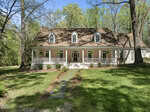 A two-story house with a tan exterior, white trim, and a wraparound front porch. The porch has hanging plants, and the house sits on a grassy lawn surrounded by tall trees. A stone path leads to the front steps.