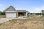 Single-story house with gray siding, a front porch, and an attached two-car garage. The yard is mostly bare with some patches of grass and straw. The house sits on a corner lot in a suburban neighborhood.