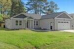 Single-story light gray house with white trim, dark gray gable accents, a covered front porch, attached two-car garage, and a well-maintained front yard with grass and young plants. Tall trees are in the background.
