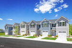 A row of modern, two-story townhouses with attached garages and small front lawns, set along a clean, paved street under a blue sky with scattered clouds.