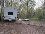 A white RV is parked next to a chain-link fence with a gate in a wooded area. A wooden deck with patio furniture sits nearby among tall trees and scattered leaves on the ground.