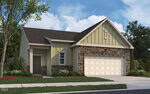 A single-story house with olive green siding, stone accents, a brown shingle roof, a white double garage door, and a small front yard with grass, bushes, and a tree. The sky is blue with scattered clouds.