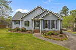 A light gray single-story house with blue shutters, a manicured lawn, flower beds, a front porch with steps, and a curved driveway sits on a sunny day at 2741 Dillon Court in Creedmoor.
