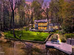 Modern house with large windows illuminated at dusk, surrounded by tall trees and lush lawn, next to a calm body of water with a dock, outdoor seating, and a swing set. The sky is a mix of purple and orange hues.