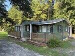 Charming small green wooden house on 1196 Cash Road in Creedmoor, featuring a covered front porch with classic white columns and dark shutters. The house sits on a grassy lot framed by trees in the background, with a gravel driveway welcoming you from the foreground.