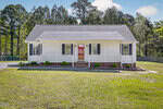 Single-story white house with a covered front porch, brick foundation, black shutters, and a gray shingled roof. The house is surrounded by grass, minimal landscaping, and trees in the background.