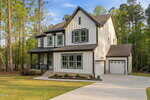 Two-story modern farmhouse with white vertical siding, black trim, covered front porch, attached two-car garage, and a neatly landscaped front yard, surrounded by tall trees.