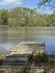 A small wooden dock extends into a calm lake surrounded by trees with green foliage near 00 Peninsula Drive in Henderson. A few houses are visible among the trees on the opposite shore under a partly cloudy sky.
