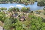 Aerial view of a lakeside neighborhood with several houses surrounded by trees, well-kept lawns, and docks along the calm water, under a partly cloudy sky.