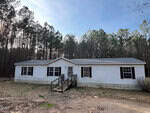 A single-story white manufactured home with black shutters, a gray shingle roof, and a central wooden ramp, set in a rural area with tall pine trees in the background under a clear sky.