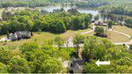 Aerial view of a suburban neighborhood with houses surrounded by trees and a large lake in the background. An arrow points to a specific house near the trees in the lower part of the image.