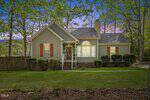 A charming light green house with white trim, brown shutters, and a front porch, set on a grassy lawn with bushes and trees. A sunset sky and wooded background complete the peaceful suburban scene.