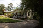 A single-story beige house with white trim and a covered front porch sits beside a curved driveway, surrounded by green grass, trees, and shrubs on a sunny day.