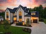 A modern two-story house with white siding, dark trim, and large windows glows warmly at sunset. Manicured lawn, young trees, and a wide driveway leading to a two-car garage complete the scene.