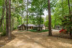 A single-story house with a stone exterior sits among tall trees, with a curved gravel driveway leading to the entrance. The surrounding area is lush and green, with a detached small building in the background.