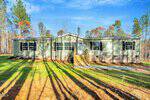 A single-story light green manufactured home with black shutters and a central front door, sits on a brick foundation surrounded by tall pine trees and a grassy yard under a blue sky.