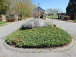 A large stone engraved with "PENINSULA" sits in the center of a landscaped roundabout at the entrance to a gated community, with brick pillars and black metal gates in the background.