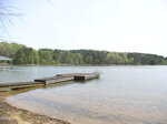 A wooden dock with a metal ladder extends into a calm lake, surrounded by trees with green foliage under a bright, clear sky. The shoreline is sandy and shallow near the dock.