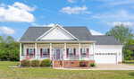 A white single-story house with a gable roof, red shutters, and a covered front porch. There is a two-car garage to the right, a brick walkway, flower beds, and a well-kept lawn under a blue sky with scattered clouds.