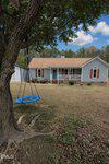 A blue circular swing hangs from a large tree in the front yard of a light gray house with brown shutters and a porch, set against a background of trees and a blue, partly cloudy sky.