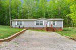 A single-story gray manufactured home with white trim sits in front of a wooded area. A gravel driveway leads up to the house, which has a small brick foundation and wooden steps up to the front door.