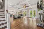 Spacious open-concept living room at 693 Hawthorne Place in Creedmoor features wooden floors, gray sectional sofa, coffered ceiling, stone fireplace, and large windows with French doors opening to a lush outdoor area. Kitchen visible in the background.