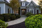 Front view of a house with a stone and gray siding exterior, large windows, wooden front door with glass panels, steps leading to the entrance, and landscaped bushes and shrubs along a curved walkway.