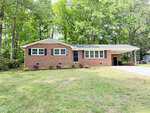 A single-story brick house with black shutters, large front windows, and a covered carport. The house is surrounded by green grass, shrubs, and tall trees in the background.
