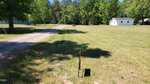 A gravel driveway curves through a grassy open yard with a small white house and trees in the background. A shadowed signpost stands in the foreground. The sky is clear and sunny.