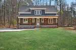 A two-story house with dark siding, white trim, and a covered front porch featuring wooden posts. The house has a gable roof, dormer windows, and is surrounded by a grassy lawn and tall trees.