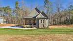A small, modern gray house with a front porch, gabled roof, and stone accents sits on a spacious lot with a fresh green lawn, concrete driveway, and surrounded by tall trees under a clear blue sky.