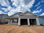 A newly built, two-story house with light gray siding, white trim, a covered front porch, and a double garage. The ground in front is bare soil, and the sky above is partly cloudy.