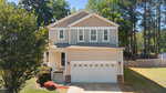 Two-story beige house with white trim and a double garage, surrounded by trees and a neatly maintained lawn. Steps lead to a small front porch, and a wooden fence runs along the right side of the backyard.