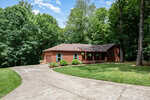 A single-story log cabin with a covered front porch and hanging plants, surrounded by trees and greenery. A curved concrete driveway leads up to the house under a partly cloudy sky.