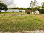 A grassy lot with a concrete slab in the foreground, bordered by trees. In the background, there is a small white shed, a tan brick building, and a large white industrial building under a cloudy sky.
