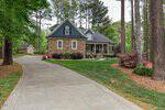A two-story house with a stone and gray exterior sits among tall pine trees, with a curved driveway, manicured lawn, shrubs, and a small shed visible in the background.