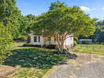 A small white house with a front porch sits under a large leafy tree, surrounded by green grass and trees. There is a gravel driveway leading up to the house and a shed in the background.