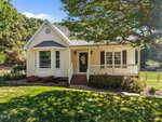 A small, single-story yellow house with white trim, a bay window, a front porch with a wreath on the door, brick steps, and a well-kept lawn, shaded by a large tree, on a sunny day.