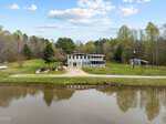 A modern white house with large windows and a screened porch sits beside a calm pond, surrounded by green trees and grass under a partly cloudy sky. A gravel driveway leads to the house.