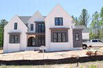 A newly constructed two-story house with white brick and gray accents, featuring large black-framed windows. A white truck is parked in the driveway, and there are construction materials and a sign in front.