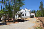 A large house under construction is surrounded by tall trees, with exterior walls covered in white building wrap. A gravel driveway leads to the garage, and a black dumpster sits at the base of the driveway.