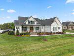 A modern white farmhouse with a dark roof, stone steps, and a covered front porch sits on a large, well-manicured lawn with young trees and shrubs, under a blue sky with scattered clouds.