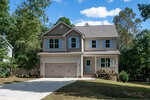 Two-story suburban house with beige siding, double garage, and gabled roof, surrounded by trees and greenery, with a concrete driveway leading up to the front. The sky is blue with scattered clouds.