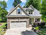 A two-story suburban house with stone and siding exterior, double garage, manicured shrubs, an American flag by the porch, and a sunny driveway, surrounded by green trees and a well-kept lawn.