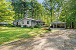 Single-story gray house with a front porch and landscaped yard, surrounded by tall trees. A detached carport with a vehicle and tools inside sits to the right on a gravel driveway. Lush green lawn and bright, sunny weather.